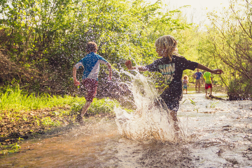 Group of children splashing in an ankle deep creek surrounded by green grass and trees on a summer day.